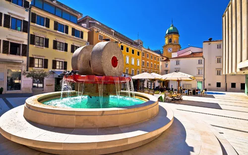 Brunnen auf einem Platz mit umliegenden historischen Gebäuden und blauem Himmel.