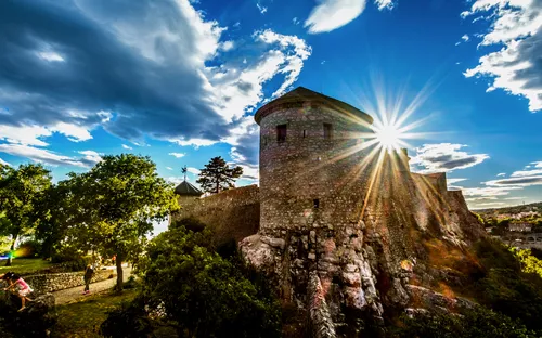 Alte Steinburg mit Sonnenstrahlen, umgeben von Bäumen und blauen Himmel.