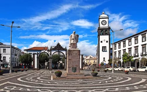 Platz mit Statue und Uhrturm, elegantes Gebäude im Hintergrund, blauer Himmel.