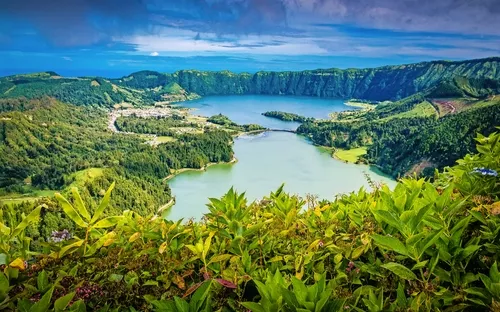 Grüner Kratersee in hügeliger Landschaft unter bewölktem Himmel.
