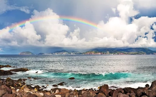 Regenbogen über Küstenlandschaft mit Meer und Wolken.