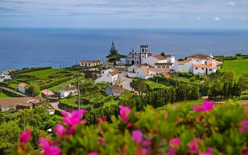 Dorf mit Kirche und Meerblick, umgeben von grüner Landschaft und Blumen im Vordergrund.