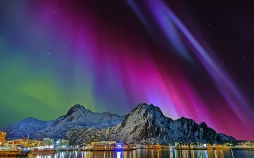 Polarlichter über schneebedeckten Bergen und einer Stadt bei Nacht.