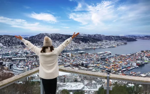 Person blickt von Aussichtspunkt auf verschneite Stadtlandschaft mit Fjord und Bergen.