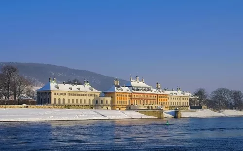 Schloss Pillnitz im Winter mit Schnee und Fluss im Vordergrund.