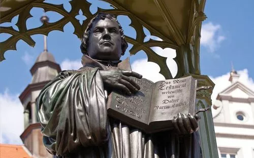 Statue eines Mannes mit offenem Buch in der Hand, historisches Gebäude im Hintergrund.
