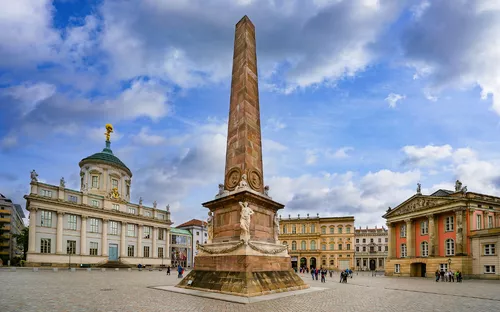 Obelisk auf einem historischen Platz mit Gebäuden im Hintergrund und bewölktem Himmel.