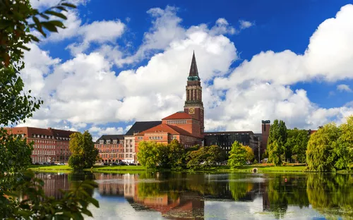 Backsteingebäude mit Turm spiegelt sich in einem See bei blauem Himmel und Wolken.