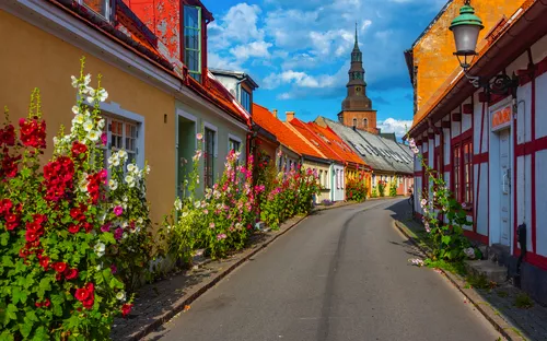 Bunte Altstadtstraße mit Blumen, Fachwerkhäuser und Kirchturm im Hintergrund bei blauem Himmel