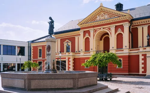 Rotes Gebäude mit Säulenfassade und Statue auf einem Brunnen im Vordergrund.
