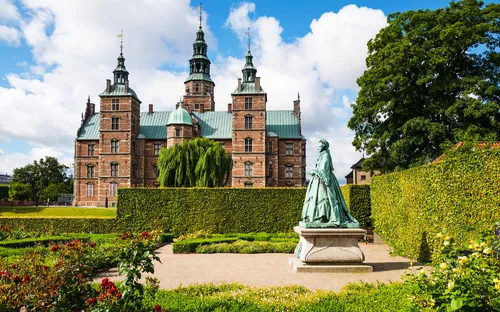 Schloss mit Türmen und Garten im Vordergrund bei blauem Himmel.