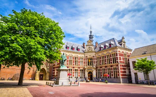 Historisches Gebäude mit Turm und Statue auf einem Platz bei sonnigem Wetter.