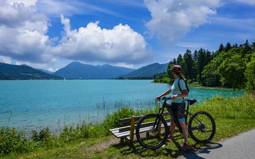 Person mit Fahrrad am See mit Bergen im Hintergrund.
