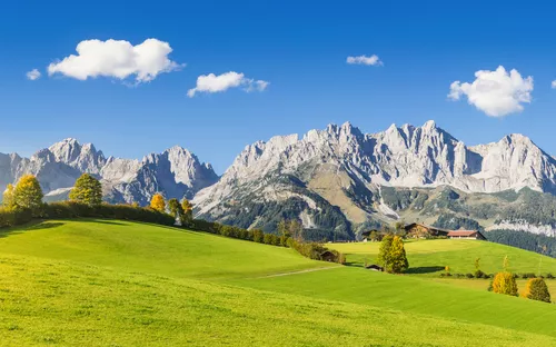 Grüne Wiesen vor einer Berglandschaft mit klarem Himmel und vereinzelten Wolken.
