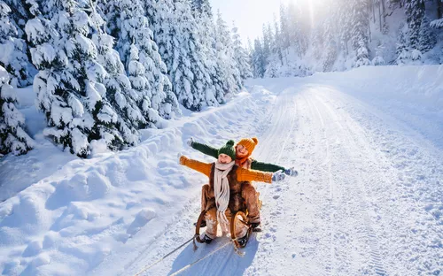 Zwei lachende Kinder rodeln im Winterwald auf einer schneebedeckten Straße.