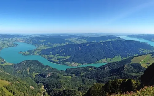 Panoramablick auf eine Landschaft mit Seen, bewaldeten Hügeln und Bergen unter blauem Himmel.