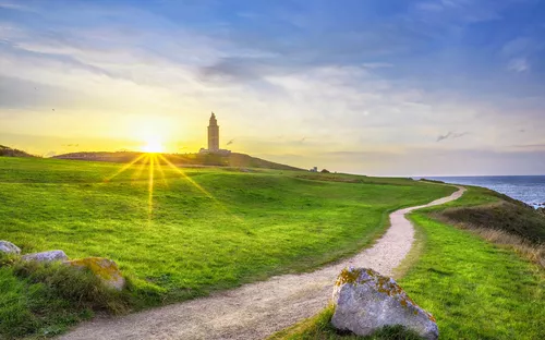 Leuchtturm am Horizont, umgeben von grünen Wiesen, bei Sonnenuntergang.