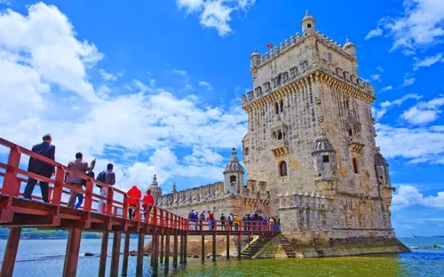Menschen auf Brücke vor dem Torre de Belém in Lissabon bei blauem Himmel.