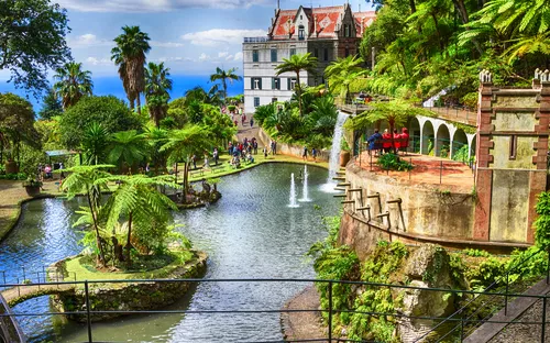 Ein tropischer Garten mit Teich, Palmen, Wasserspielen und historischem Gebäude im Hintergrund.