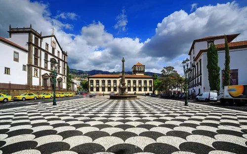 Schachbrettartiger Platz mit Brunnen, umgeben von historischen Gebäuden und Bergen im Hintergrund.