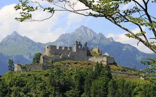 Burg auf einem bewaldeten Hügel vor alpenländischer Bergkulisse.