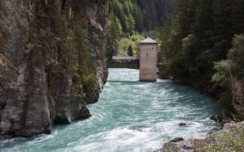 Fluss in Gebirgsschlucht mit Turm und Brücke, umgeben von Bäumen.