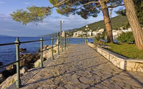Steinpflasterweg entlang der Küste mit Bäumen und Stadt im Hintergrund.