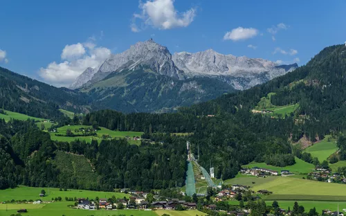 Dorf in alpiner Landschaft mit Bergen und grünen Feldern bei klarem Himmel.