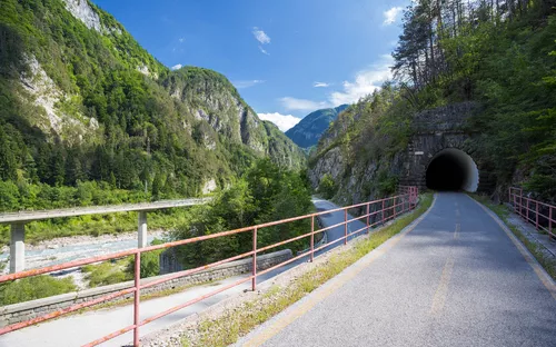 Straße mit Tunnel in gebirgiger Landschaft, umgeben von grünen Bäumen und Fluss.