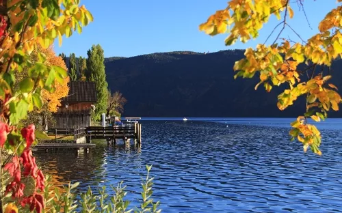 See mit Herbstbäumen, Bootsanleger und bewaldeten Hügeln im Hintergrund.
