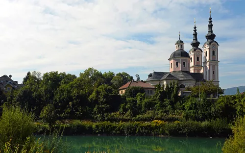 Barockkirche mit Türmen am Flussufer, umgeben von Bäumen und bewölktem Himmel.