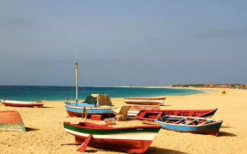 Bunte Boote stehen auf einem sandigen Strand mit blauem Meer im Hintergrund.