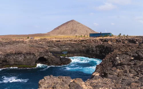 Felsige Küste mit blauem Meer und Vulkan im Hintergrund.