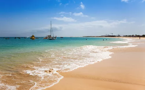 Strand mit blauem Himmel, sanften Wellen und Booten im Wasser.