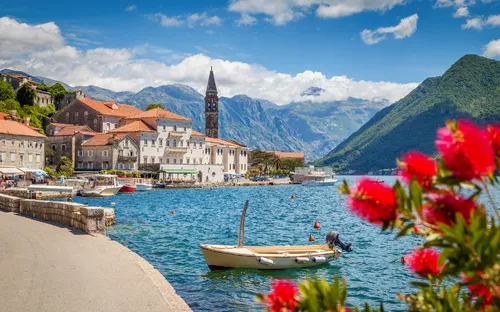 Küstenstadt mit Glockenturm am blauen Meer, umgeben von Bergen und blühenden Blumen.