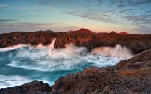 Steilküste mit brechenden Wellen bei Sonnenuntergang, im Hintergrund Berge.