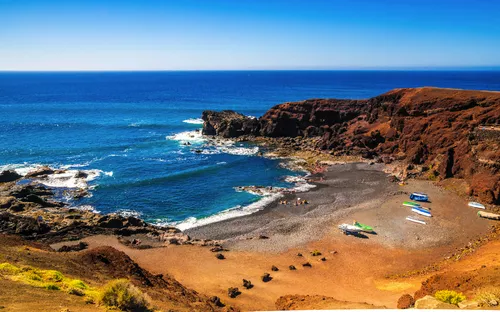 Kleine Bucht mit schwarzem Sandstrand und blauem Meer, umgeben von Felsen.