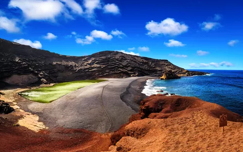Küstenlandschaft mit grünem See, schwarzem Strand und blauem Himmel.