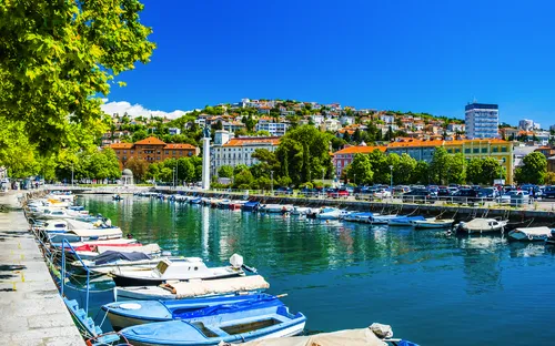 Uferpromenade mit Booten, blauen Himmel, und Stadt im Hintergrund.