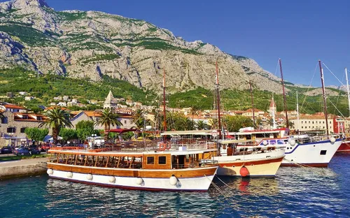 Boote liegen im Hafen vor einer Stadt am Fuße eines großen Gebirges mit grüner Vegetation.