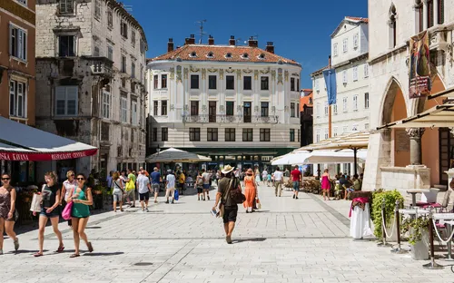 Menschen spazieren auf einem sonnigen Platz umgeben von historischen Gebäuden.