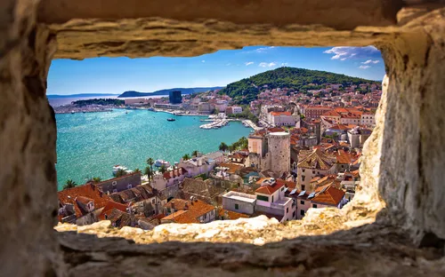 Blick durch Steinfenster auf Küstenstadt mit Meer und Hügel im Hintergrund.