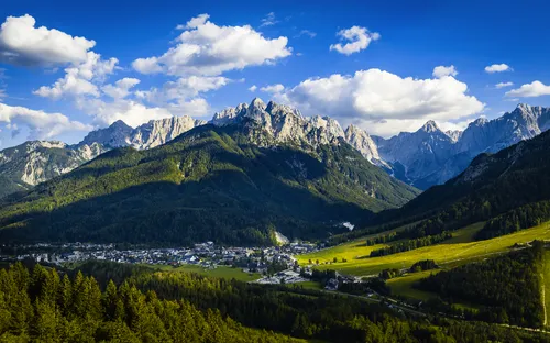 Berglandschaft mit blauem Himmel und kleinen Wolken über einem Tal mit Dorf und Wäldern.