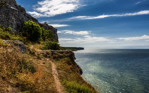 Küste mit Felsen und Wanderpfad, Meer im Hintergrund bei blauem Himmel.