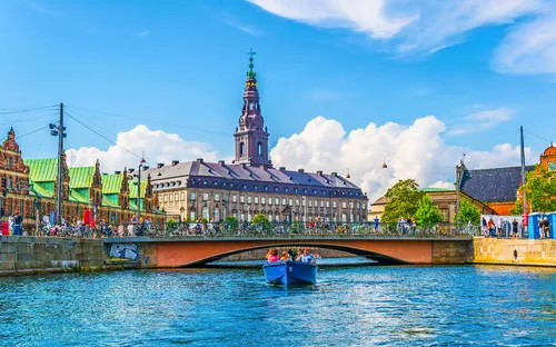 Menschen in einem blauen Boot auf einem Kanal vor historischem Gebäude an einem sonnigen Tag.