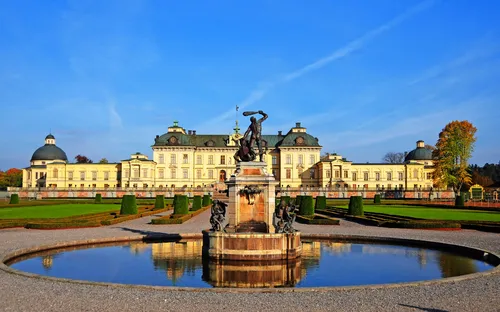 Barockschloss mit Park und Springbrunnen im Vordergrund bei sonnigem Wetter.