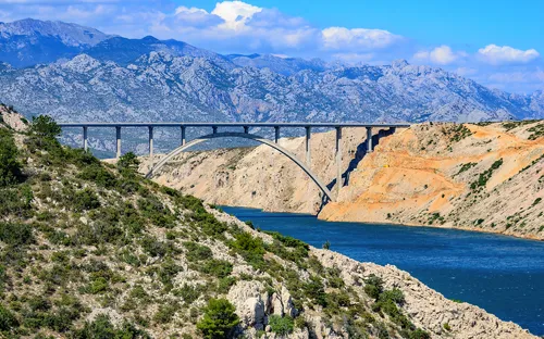 Brücke über Fluss zwischen felsigen Hügeln und Bergen im Hintergrund.