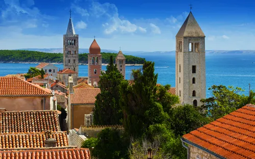 Mittelalterliche Stadt mit Kirchtürmen und Meer im Hintergrund bei klarem Himmel.