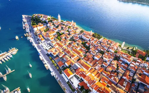 Luftaufnahme einer Küstenstadt mit roten Dächern und Hafen am Meer.