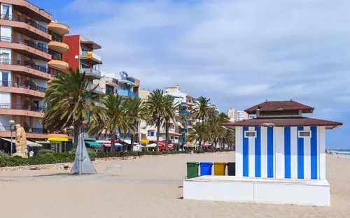 Strand mit blau-weißem Kiosk, Palmen und Wohnhäusern im Hintergrund.